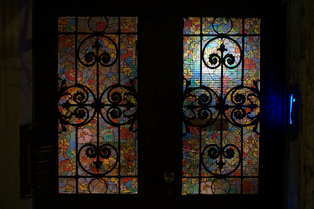 A house door with very colourful ornamental windows in it. On the side there's an intercom with bright blue light.